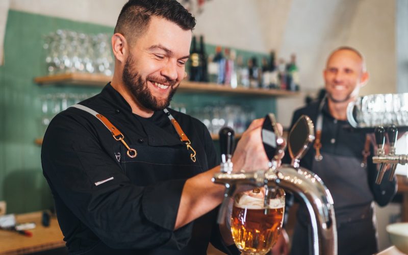 A man pours beer into a glass from a tap. He's smiling and wearing a black apron. Another man is nearby.