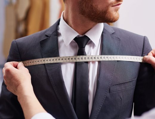 A man wearing a tuxedo jacket and a white shirt stands as a tailor stretches a measuring tape over the man's chest.