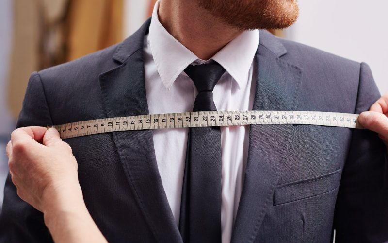 A man wearing a tuxedo jacket and a white shirt stands as a tailor stretches a measuring tape over the man's chest.