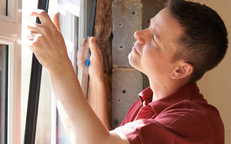 A man carefully installs a replacement window frame, aligning the frame and glass to a partially constructed wall.