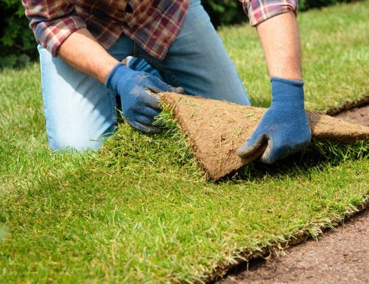 A man kneeling and installing a turf patch in a backyard. He wears blue gloves, jeans, and a plaid shirt.