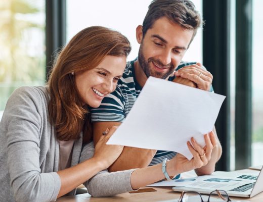 A man and woman sitting at a table with a laptop, glasses, and documents, smiling and looking at a piece of paper.