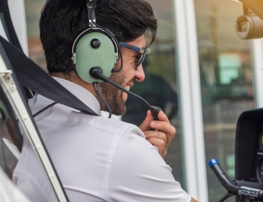 A smiling pilot wearing a headset and sunglasses talks into his radio while sitting in the cockpit of a small plane.