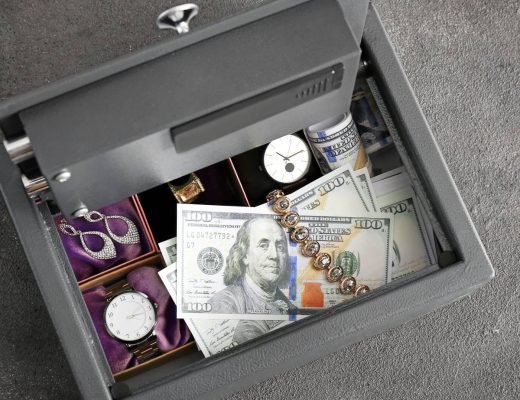 An open safe sits on gray flooring. The safe's compartments contain jewelry, watches, and hundred-dollar bills.