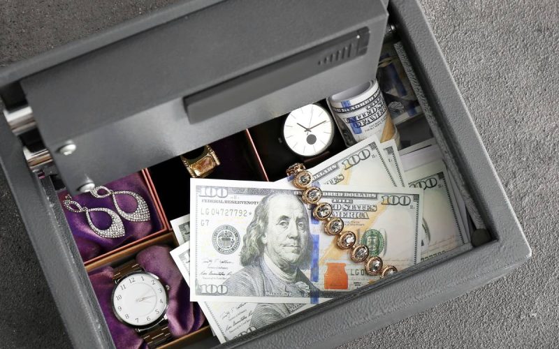 An open safe sits on gray flooring. The safe's compartments contain jewelry, watches, and hundred-dollar bills.