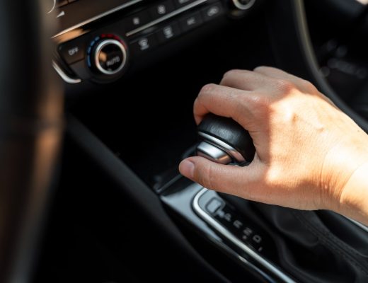 A close-up view shows a driver resting their hand on a black gear shifter with a silver button on the side.