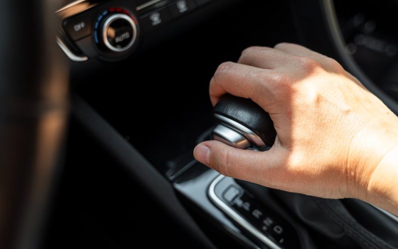 A close-up view shows a driver resting their hand on a black gear shifter with a silver button on the side.