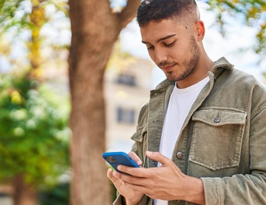 A man in a white shirt and green jacket looking at his cell phone while standing outside. The phone has a blue case.