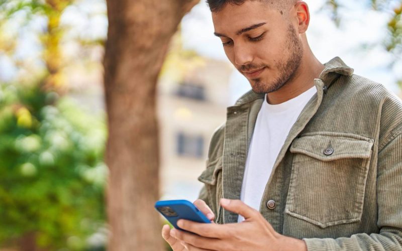 A man in a white shirt and green jacket looking at his cell phone while standing outside. The phone has a blue case.