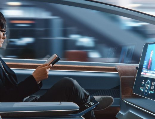 A man sits in the front seat of an autonomous vehicle, reading a book. The vehicle contains a large screen with a person on it.