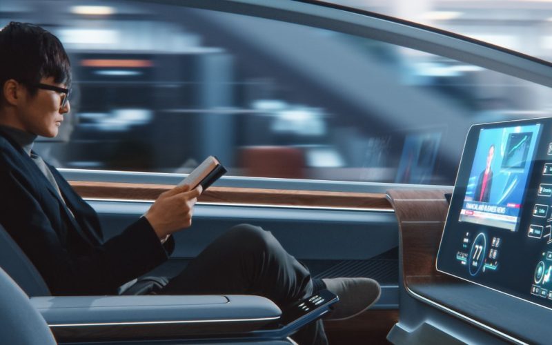 A man sits in the front seat of an autonomous vehicle, reading a book. The vehicle contains a large screen with a person on it.