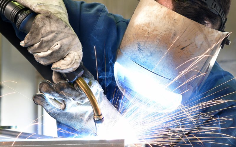 A man wearing a protective visor is using a torch to weld a metal piece in a garage, sparks flying from the worksite.