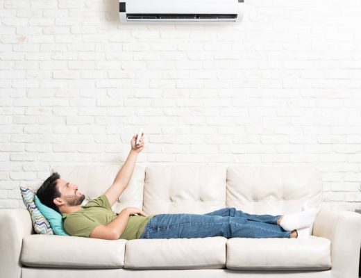 A man lying on a white couch in a living room and pointing a remote control at a mini split on the white-brick wall above him.