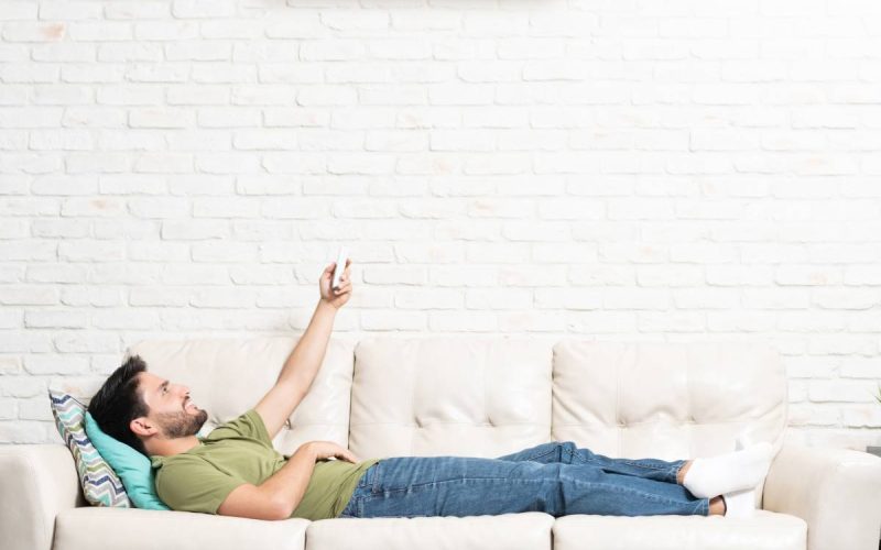 A man lying on a white couch in a living room and pointing a remote control at a mini split on the white-brick wall above him.