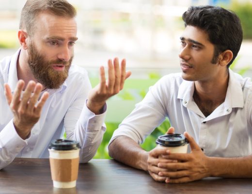 Two men sit at a wooden table outside with to-go coffee cups. One man has his arms up, and the other is listening.