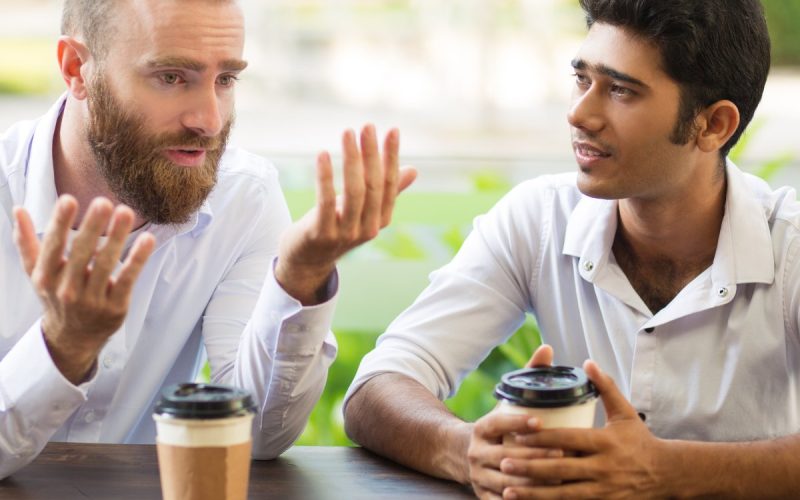 Two men sit at a wooden table outside with to-go coffee cups. One man has his arms up, and the other is listening.