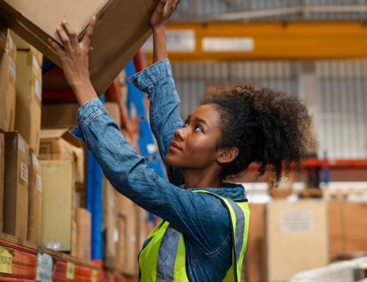 A woman wearing a blue shirt and bright yellow safety vest puts a cardboard box up on a warehouse shelf.