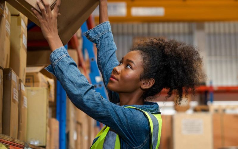 A woman wearing a blue shirt and bright yellow safety vest puts a cardboard box up on a warehouse shelf.