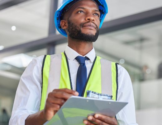 A black man wearing a neon yellow and grey safety vest and a blue hard hat holds a clipboard in his hands.