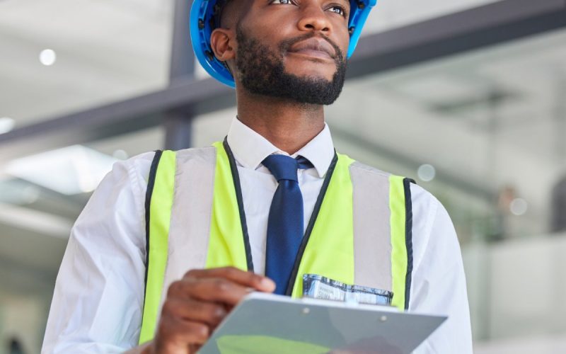 A black man wearing a neon yellow and grey safety vest and a blue hard hat holds a clipboard in his hands.