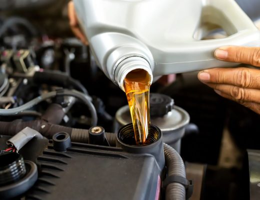 An auto mechanic pouring a bottle of engine oil into a car's oil reservoir. The engine oil is light brown in color.