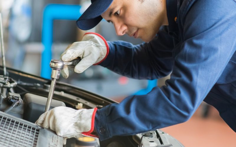 A man in blue full-body work outfit, blue baseball hat, and gloves uses a ratchet wrench to work under a car hood.