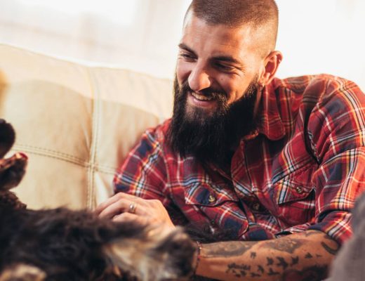 A happy man sits on a couch and pats his dog. The man has a beard and arm tattoos, and the dog is black.