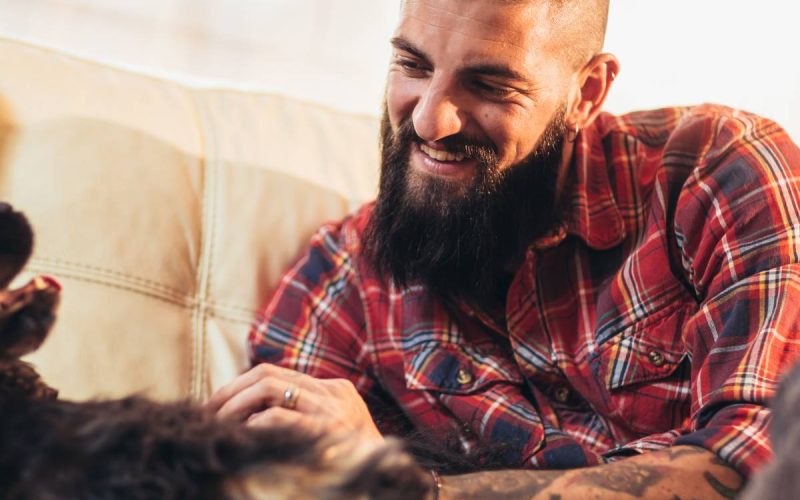 A happy man sits on a couch and pats his dog. The man has a beard and arm tattoos, and the dog is black.