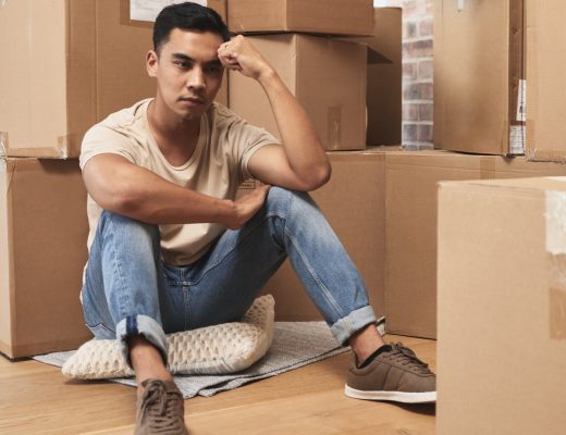 A man sitting on his hardwood floor surrounded by cardboard boxes appearing stressed with his head in his hand.