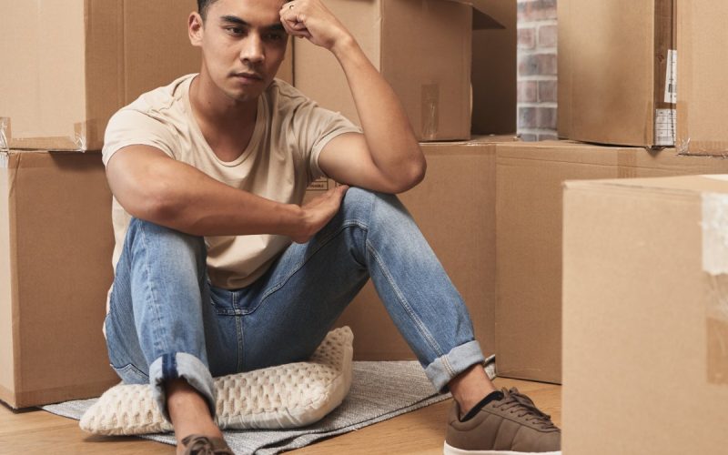 A man sitting on his hardwood floor surrounded by cardboard boxes appearing stressed with his head in his hand.