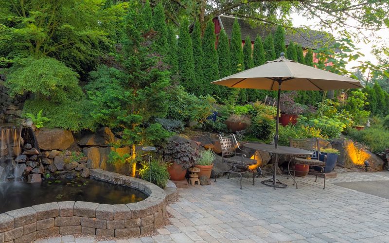 A backyard patio with a gray brick foundation, a decorative fountain, and a table with two chairs and an umbrella.