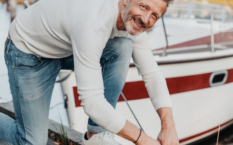 An older man kneeling on a dock tying a dock line from his boat that is floating in the marine behind him.