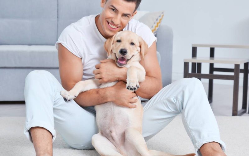 A man sits on the floor with a labrador retriever puppy between his legs. They're both smiling and looking at the camera.