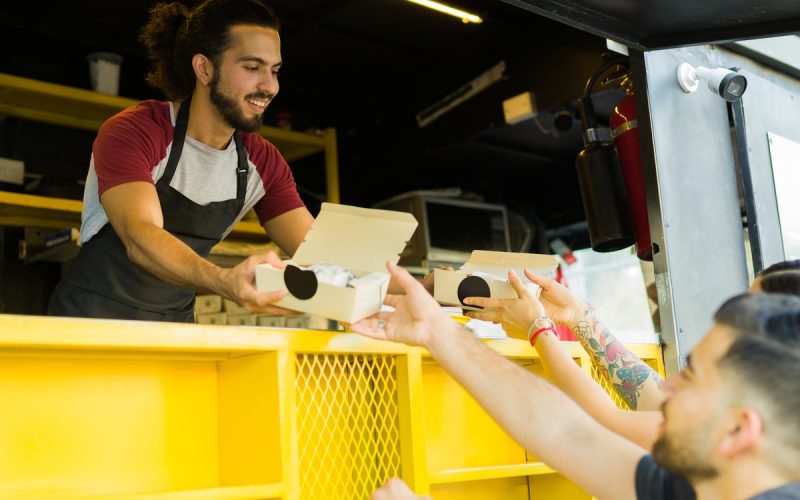 A smiling man running a food truck hands out boxed sandwiches to two customers standing at the open window.