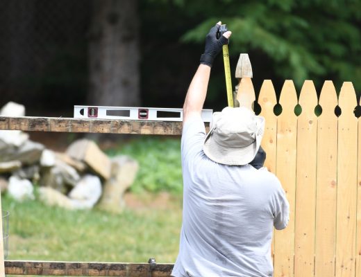 A rear view of a man measuring a wood panel with measuring tape as he installs a wooden fence in a home backyard.