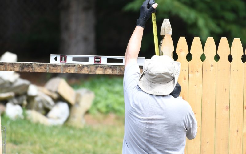 A rear view of a man measuring a wood panel with measuring tape as he installs a wooden fence in a home backyard.