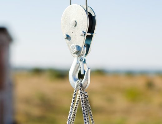 A closeup of a hook attached to a rope and cables lifting an object on a construction site in front of a blurry background.