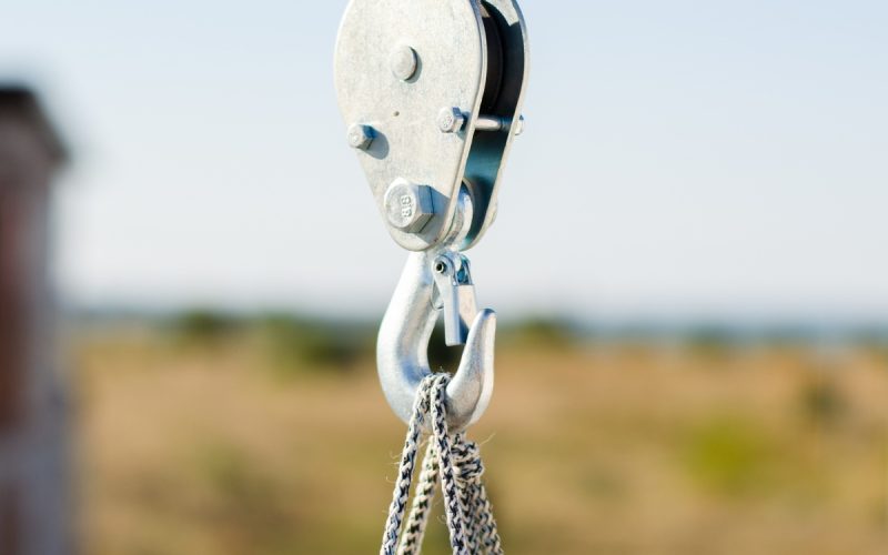 A closeup of a hook attached to a rope and cables lifting an object on a construction site in front of a blurry background.