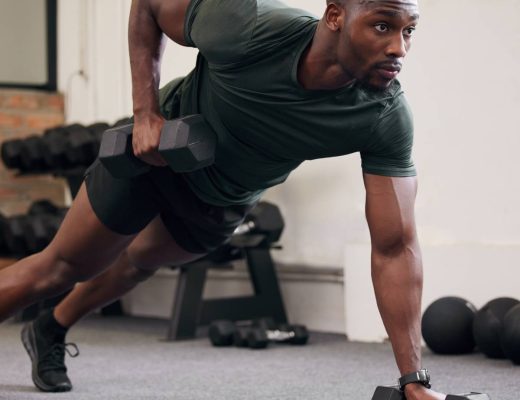 A man wearing dark shorts and a T-shirt using two black dumbbells to hold his body in a plank position at a gym.