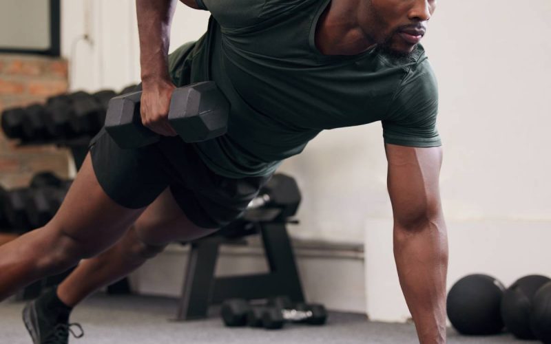 A man wearing dark shorts and a T-shirt using two black dumbbells to hold his body in a plank position at a gym.