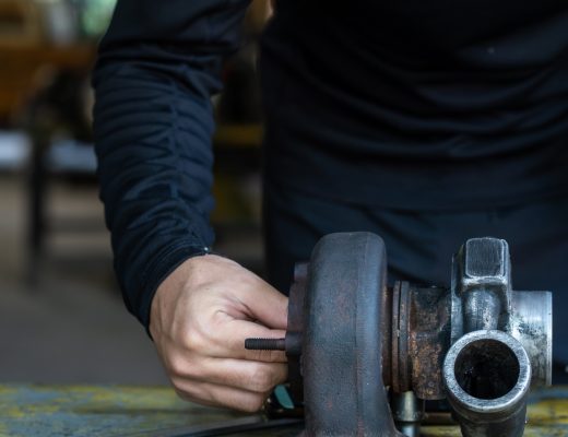 A worker in black repairs a turbocharger with a wrench at a worn workbench with tools in an industrial setting.