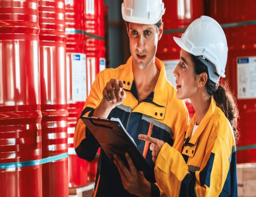 A woman and man in hardhats and safety gear stand in a warehouse filed with barrels and discuss a document.