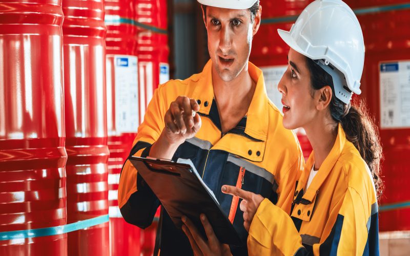 A woman and man in hardhats and safety gear stand in a warehouse filed with barrels and discuss a document.