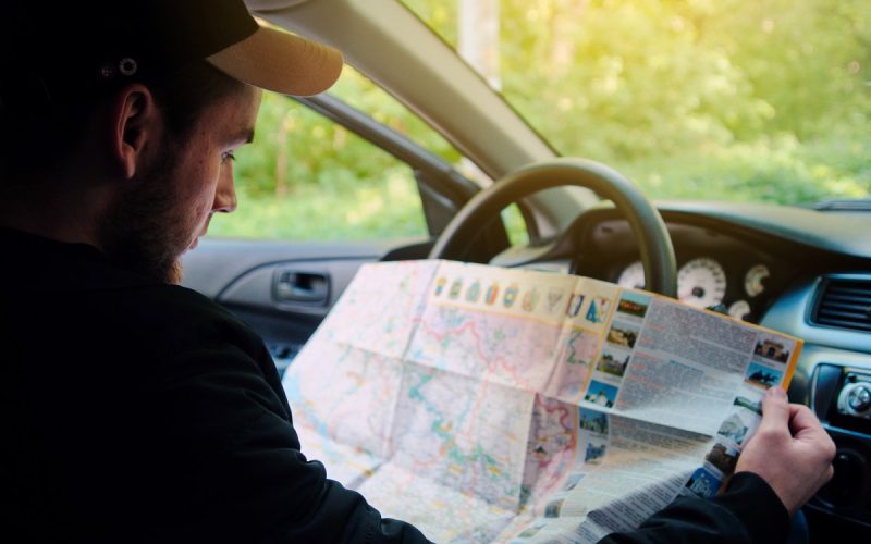 A man in a baseball cap with a yellow brim reading a paper map while sitting behind the wheel of his car.