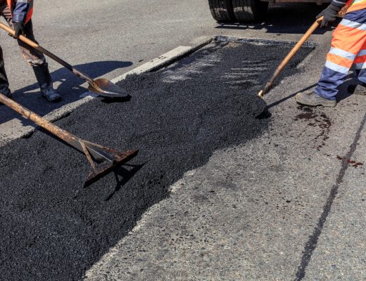 Three asphalt repair employees in orange safety vests use shovels to disperse asphalt pavement on the road.