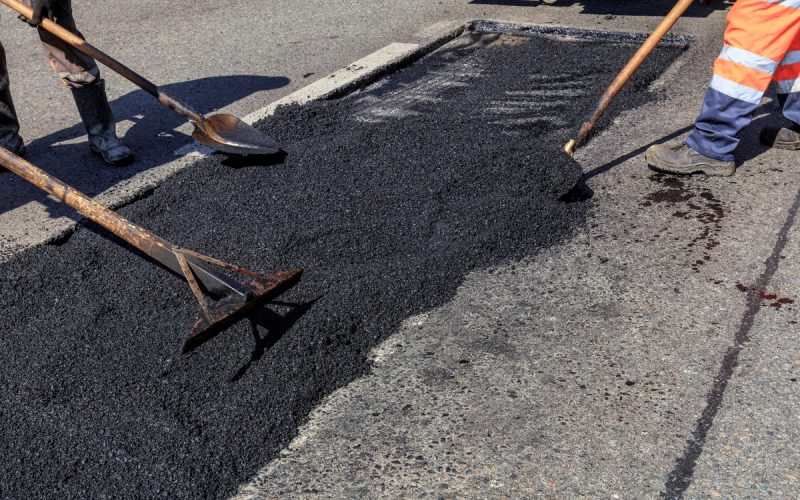 Three asphalt repair employees in orange safety vests use shovels to disperse asphalt pavement on the road.