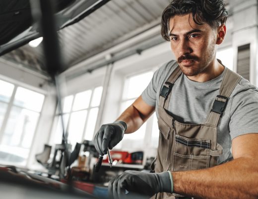A young man in a garage is working on the engine of a car. He is wearing brown overalls and gray gloves.
