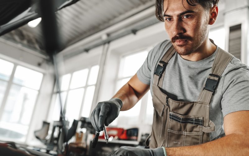 A young man in a garage is working on the engine of a car. He is wearing brown overalls and gray gloves.