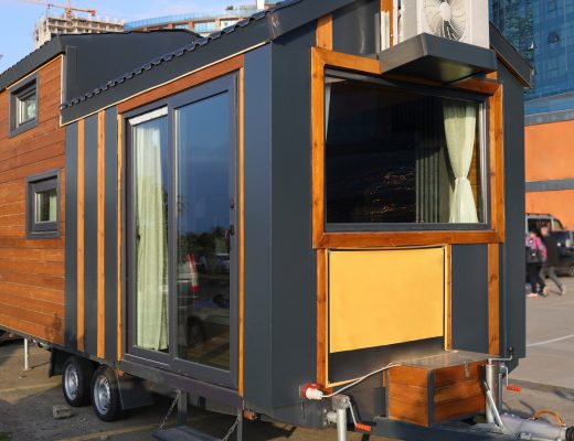 A small, wooden mobile home with black accent colors sits parked on its wheels in a parking lot. Cars are parked nearby.