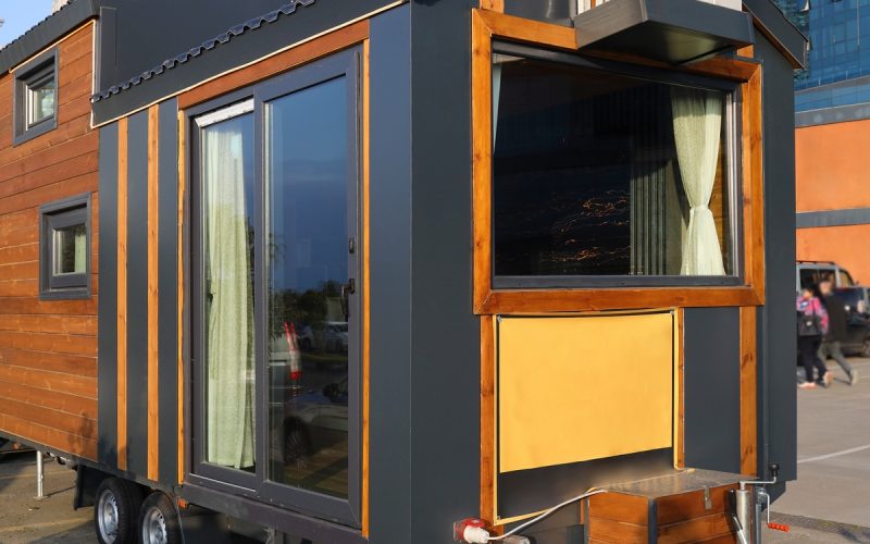 A small, wooden mobile home with black accent colors sits parked on its wheels in a parking lot. Cars are parked nearby.